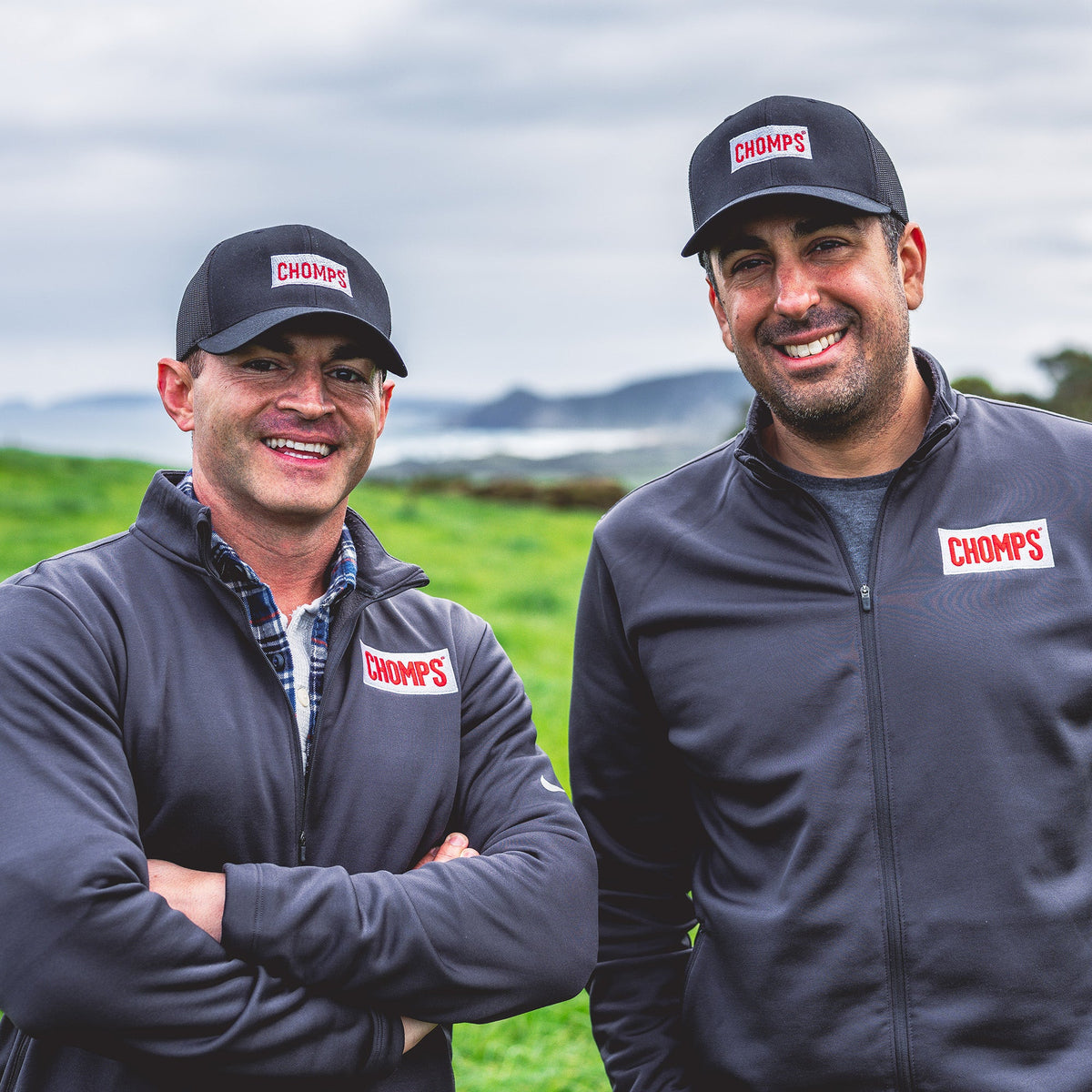 Pete and Rashid standing outdoors in a grassy field under an overcast sky, wearing matching dark gray CHOMPS branded zip-up jackets and baseball caps.
